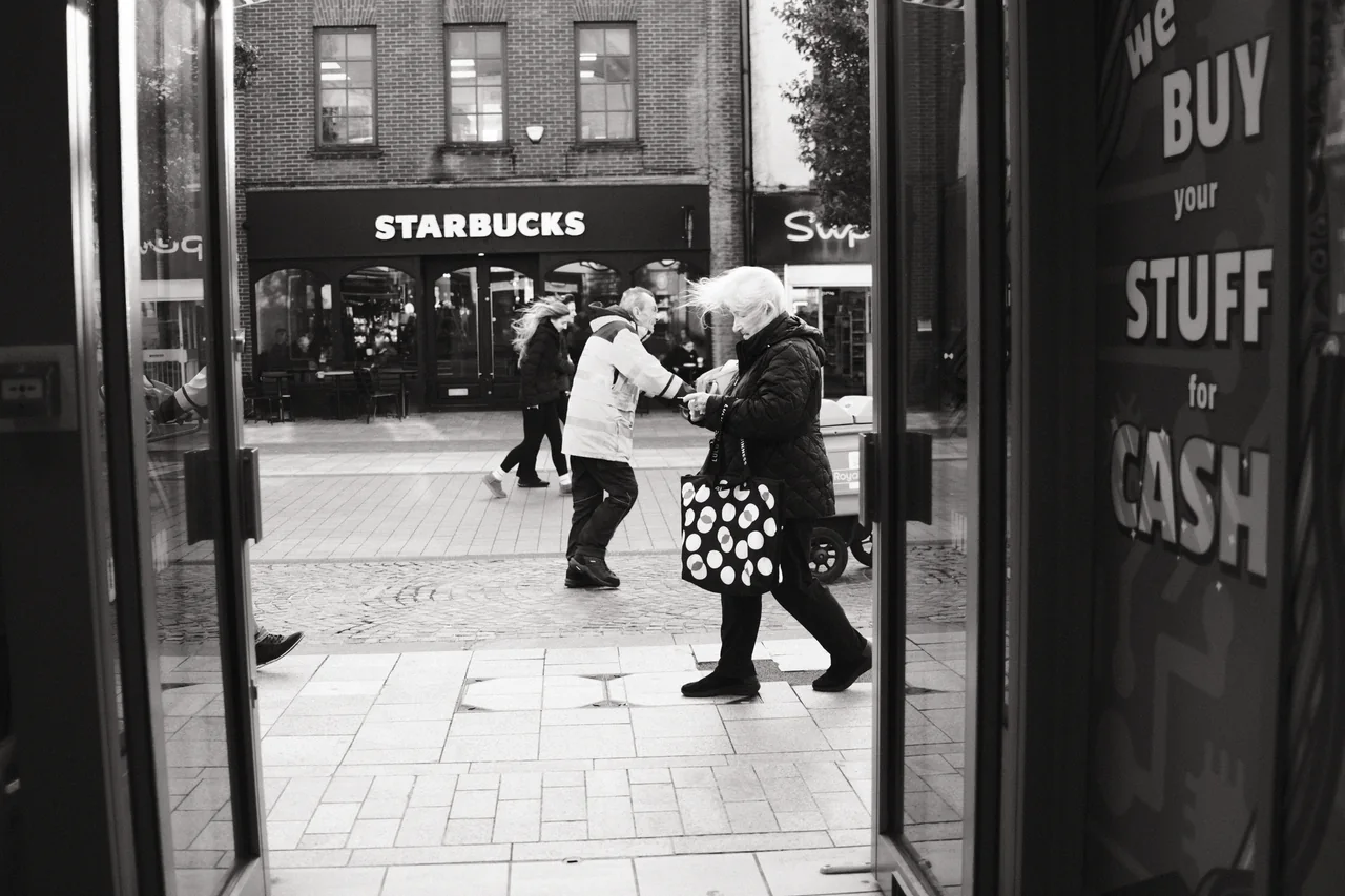 Seen through an open shop doorway, a woman carrying a spotted bag passes a postal worker and other pedestrians. Black and white photograph by Mark C Grant.