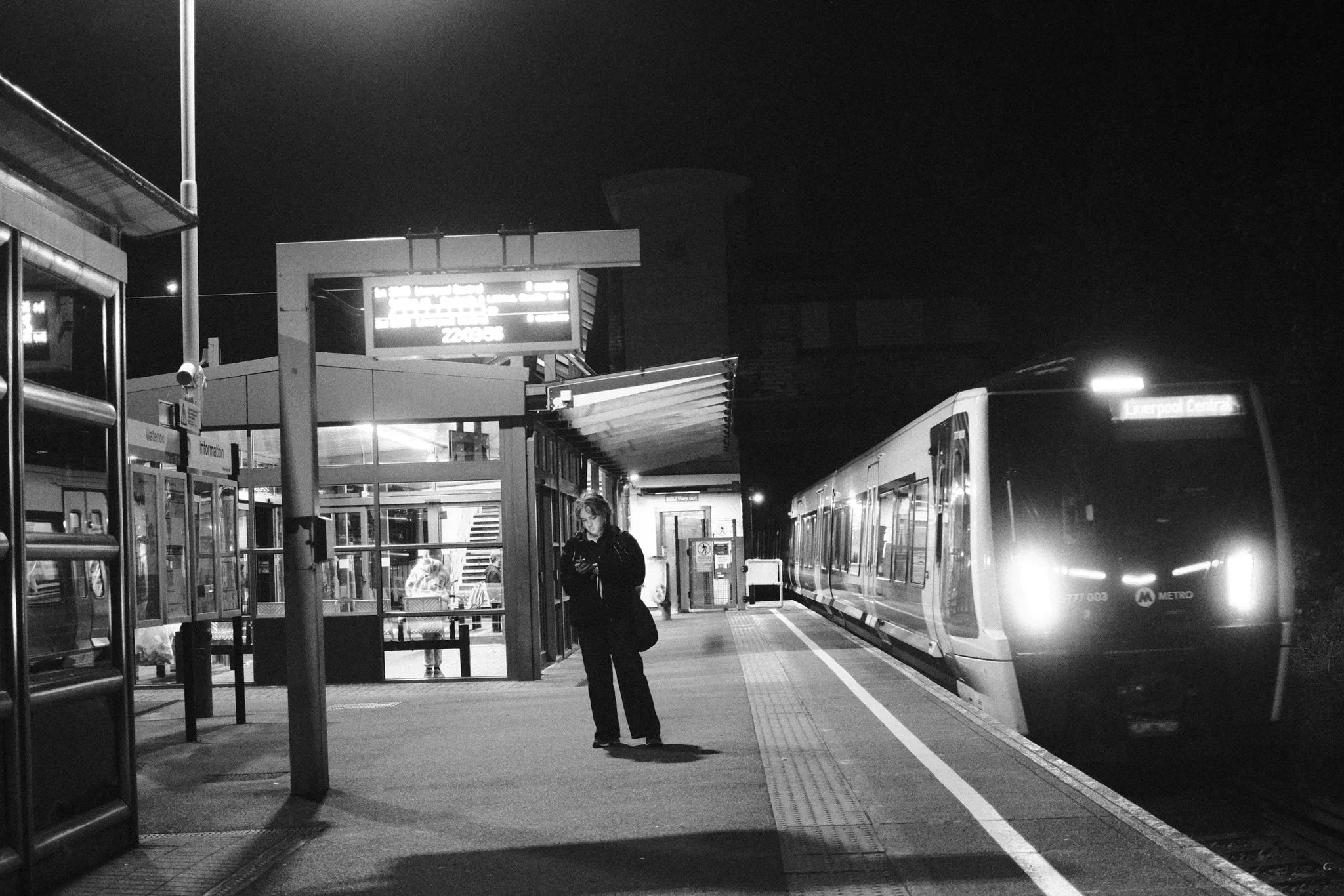 A train with bright headlights approaching a person on the platform. Black and white photograph by Mark C Grant.