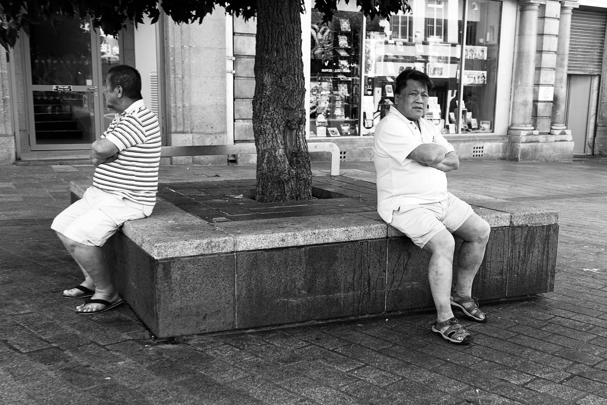 Two people seated on opposite ends of a raised tree surround, in black and white. Photograph by Mark C Grant.