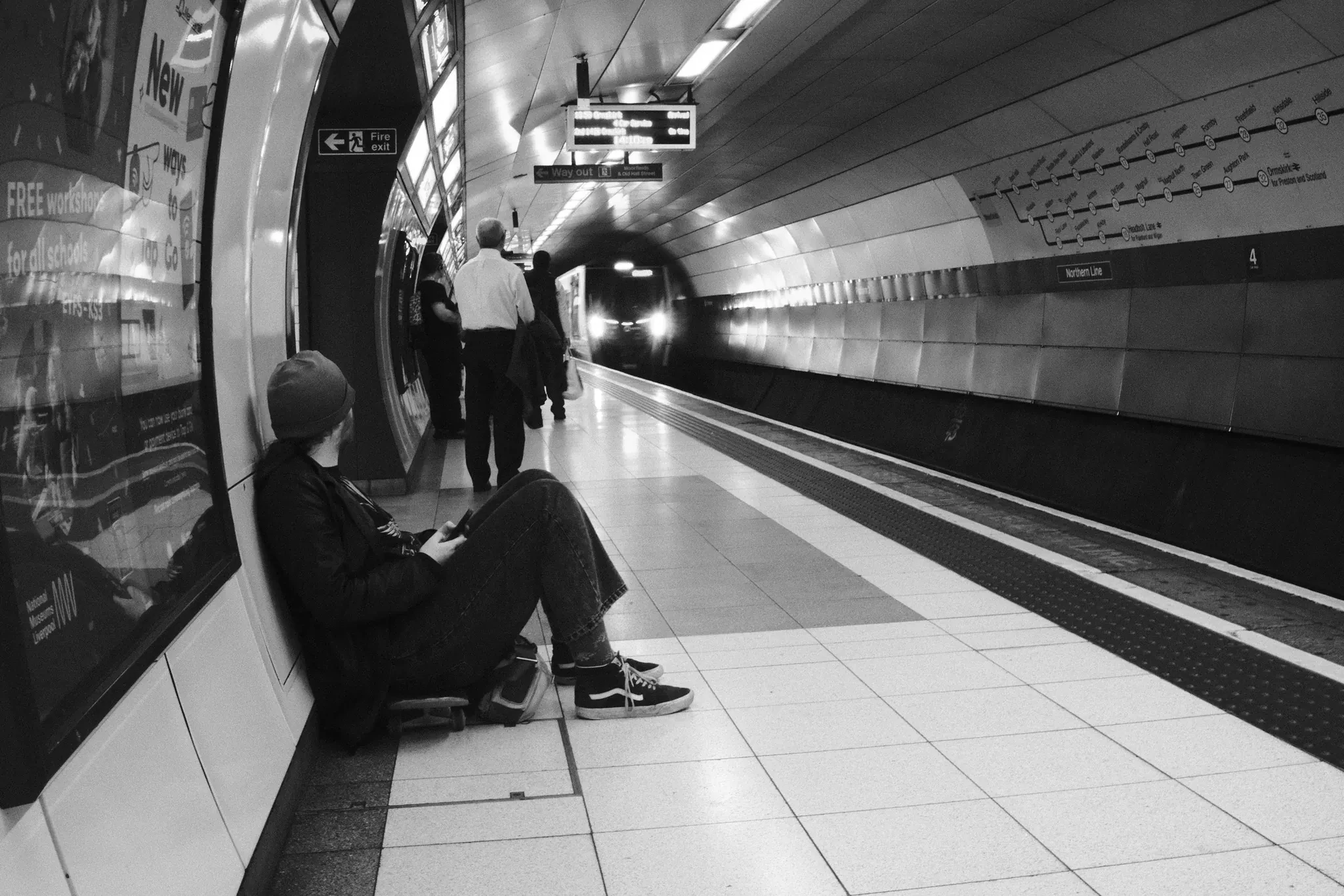 A seated passenger waits on a Moorfields platform as a train approaches through the tunnel. Black and white photograph by Mark C Grant.