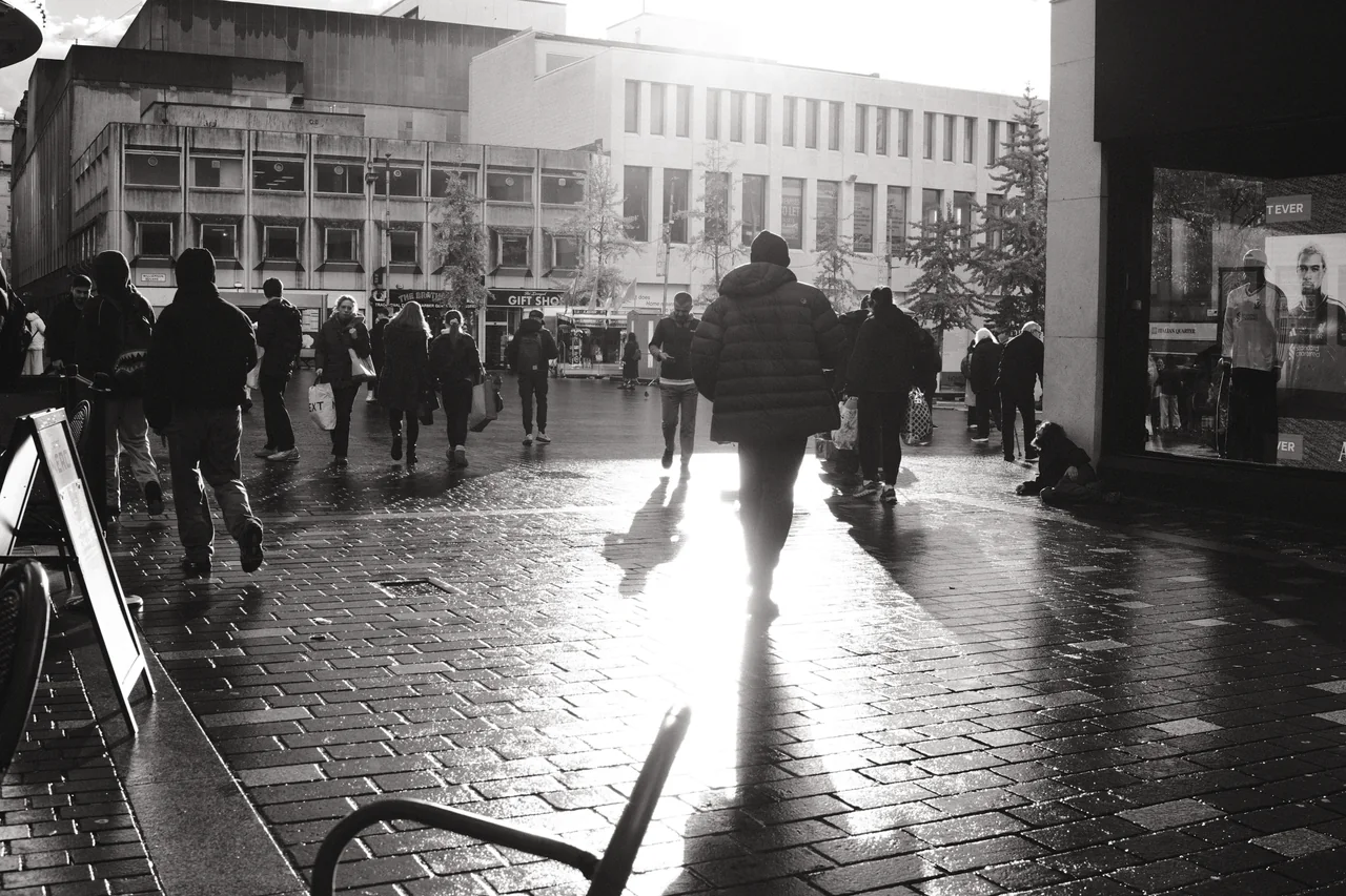 A hooded figure walks into reflected sunlight on the wet paving of Williamson Square, Liverpool. Black and white photograph by Mark C Grant.