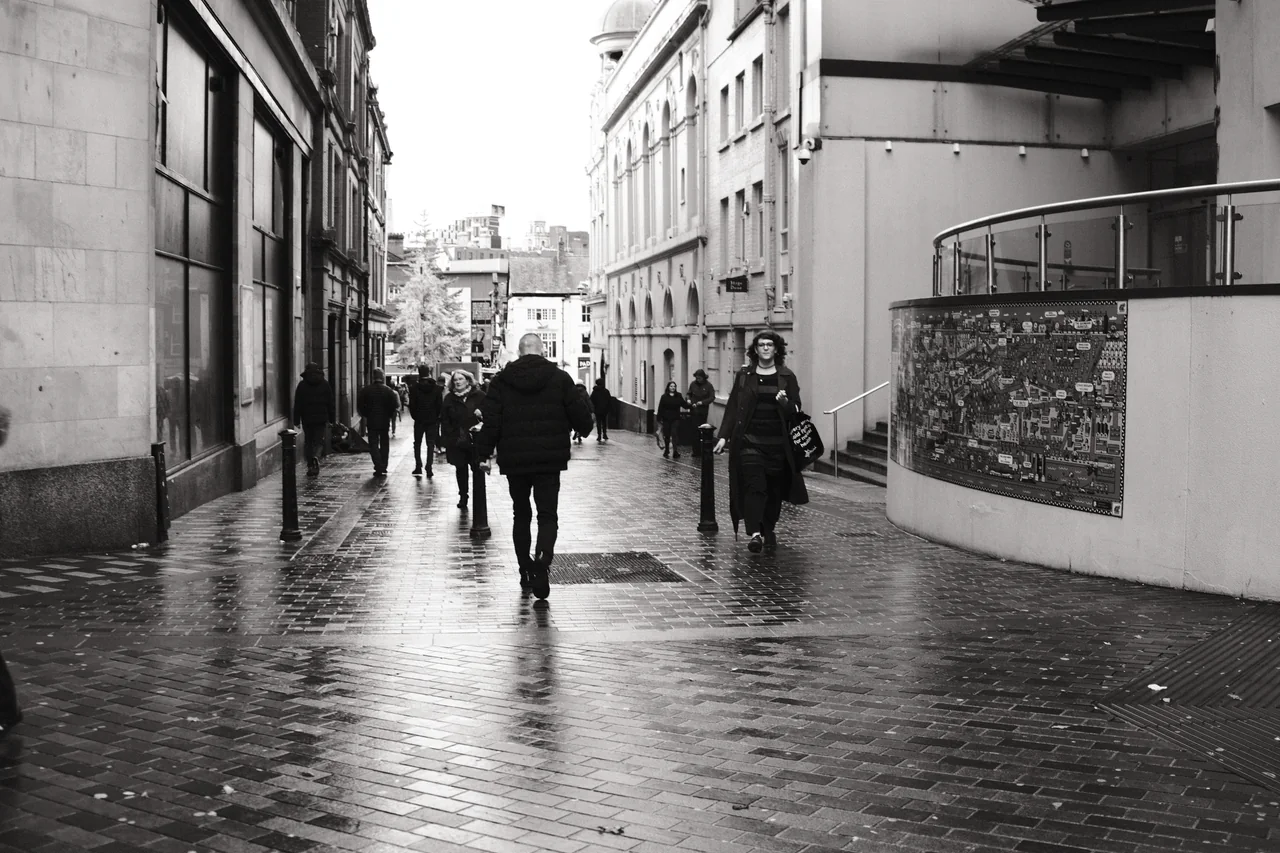 Pedestrians move along reflective paving beside Liverpool's Playhouse Theatre, with its stage door on the right. Black and white photograph by Mark C Grant.